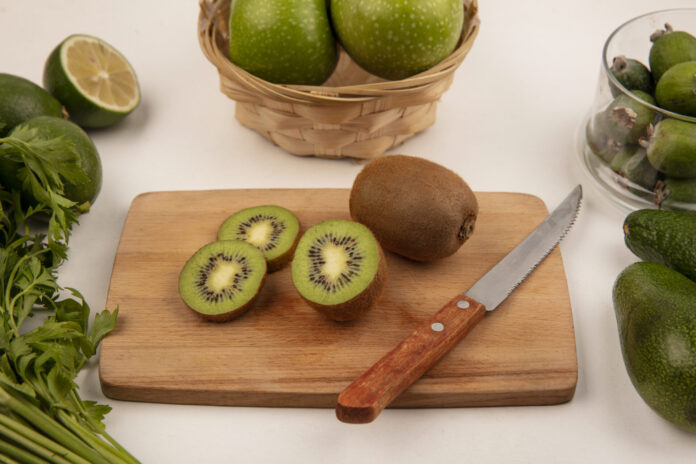 top view of fresh kiwis on a kitchen board with knife with apples on a bucket with feijoas on a glass bowl with limes and avocados isolated on a white background